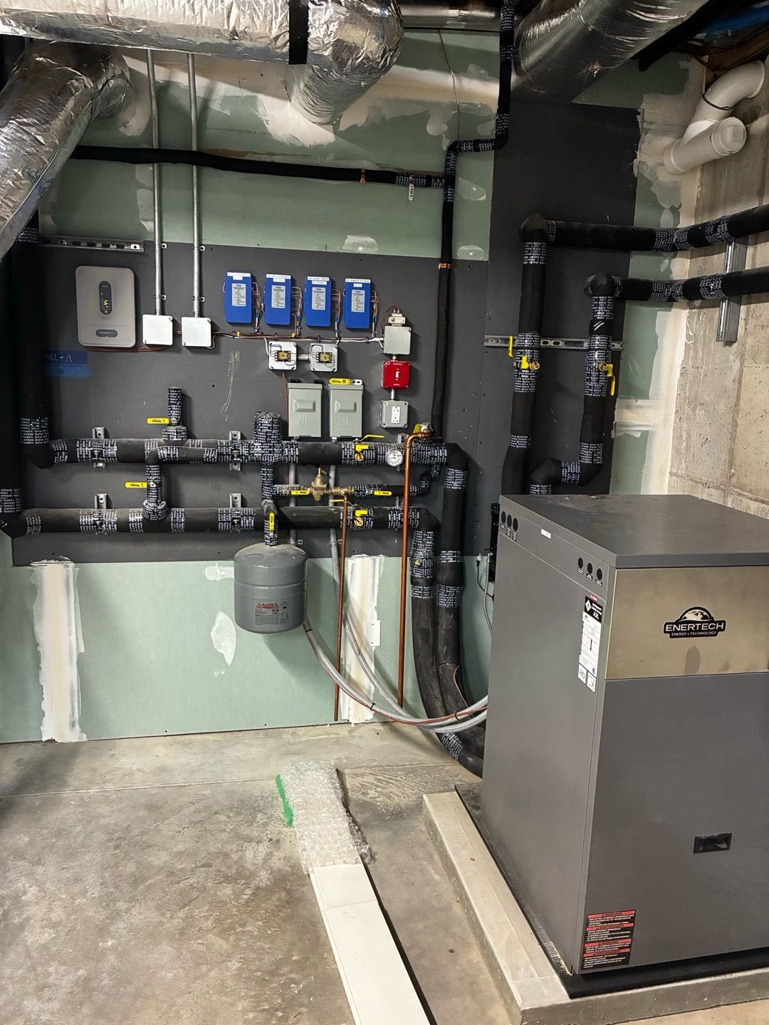 Mechanical room with pipes, valves, blue controls, and a large HVAC unit against concrete walls.