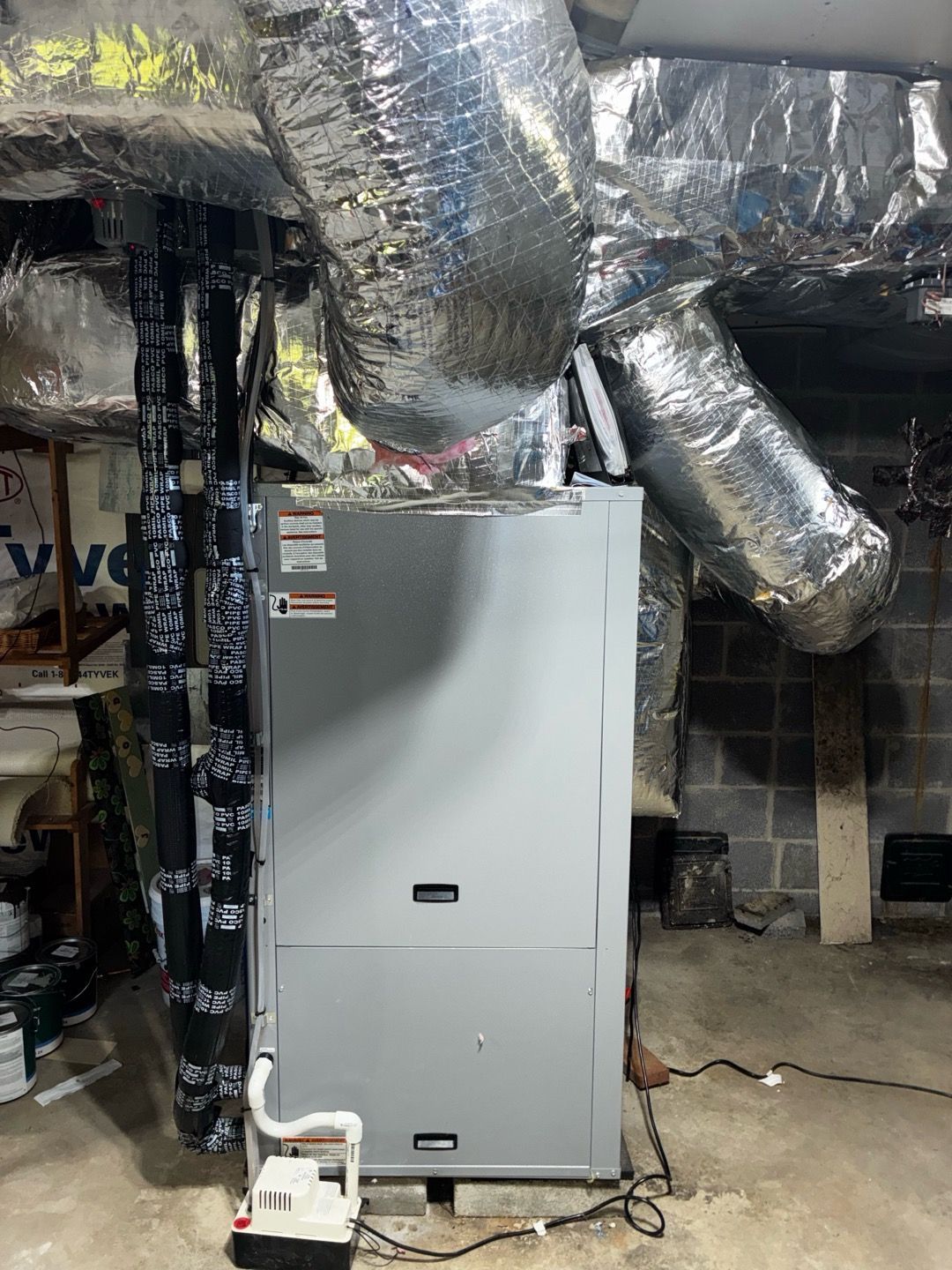 Gray furnace in unfinished basement with exposed silver ductwork and a white portable unit on the floor