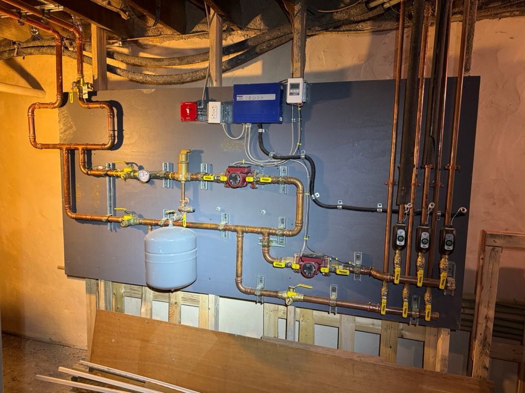 Basement utility wall with pipes, valves, gauges, and a blue water tank under exposed wiring