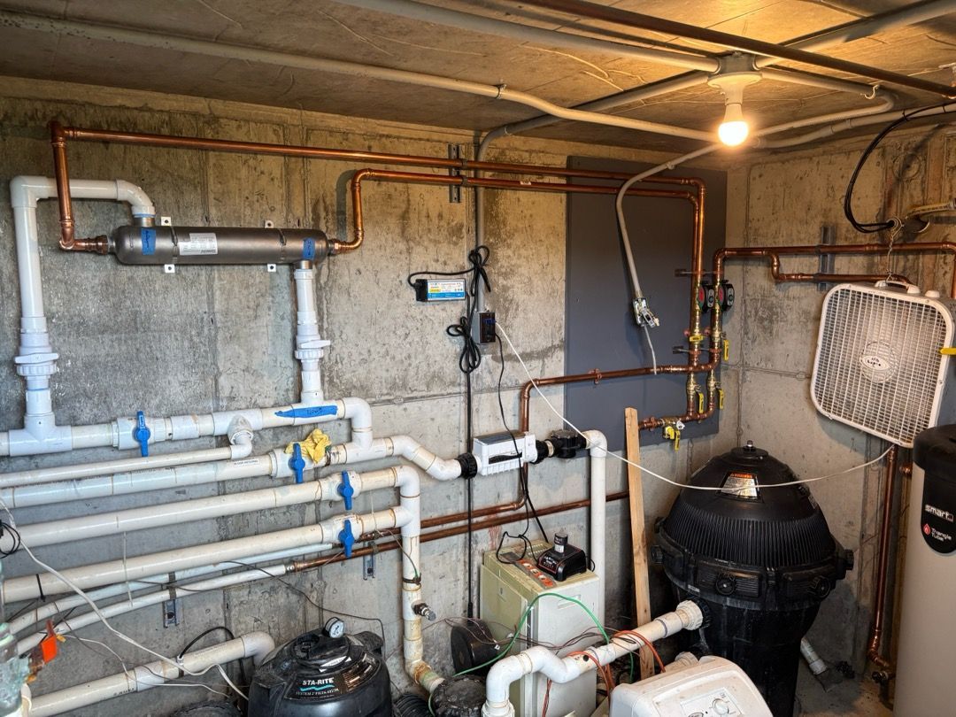 Basement utility room with exposed pipes, valves, water heater, and a fan on the wall.