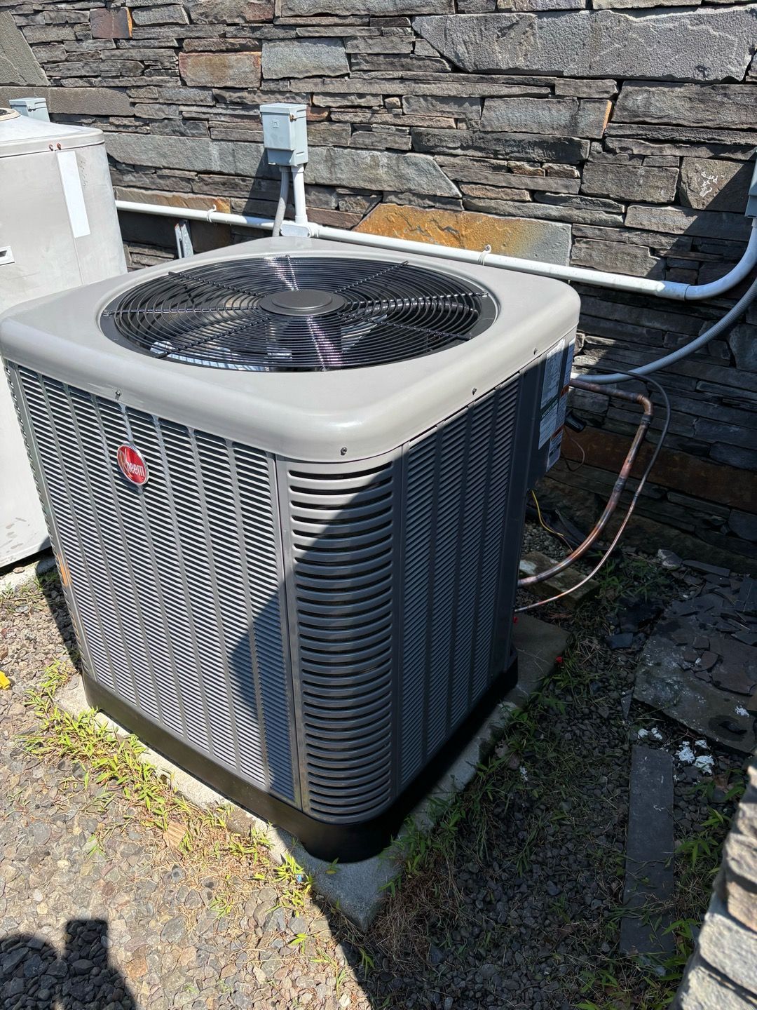 Outdoor HVAC condenser unit beside a stone wall, with a fan on top and connected copper and electrical lines.