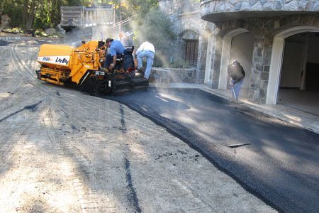 Road paving crew laying fresh asphalt beside a garage in a wooded area