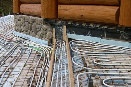 Coiled white tubing laid beneath a log cabin wall and stone foundation during installation.