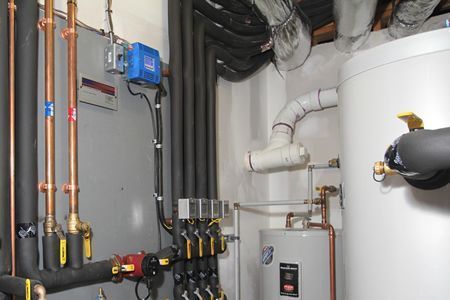 Mechanical room with pipes, valves, pumps, and a white tank against the wall