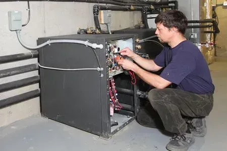 Technician crouches beside an open HVAC unit, adjusting wiring in a basement utility room