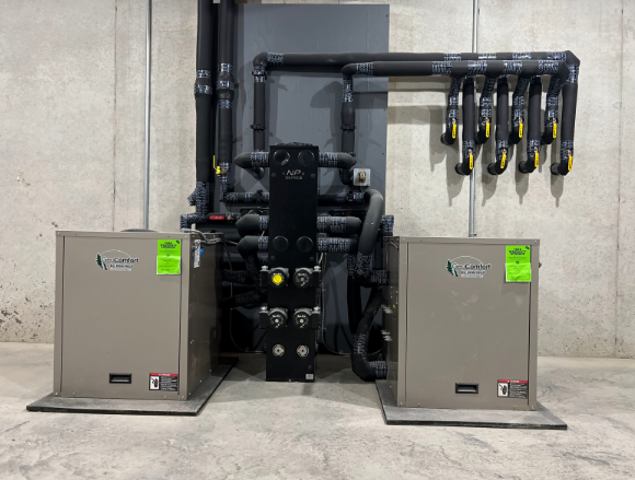 Industrial HVAC equipment with two gray units, black piping, and a central pump in a basement room