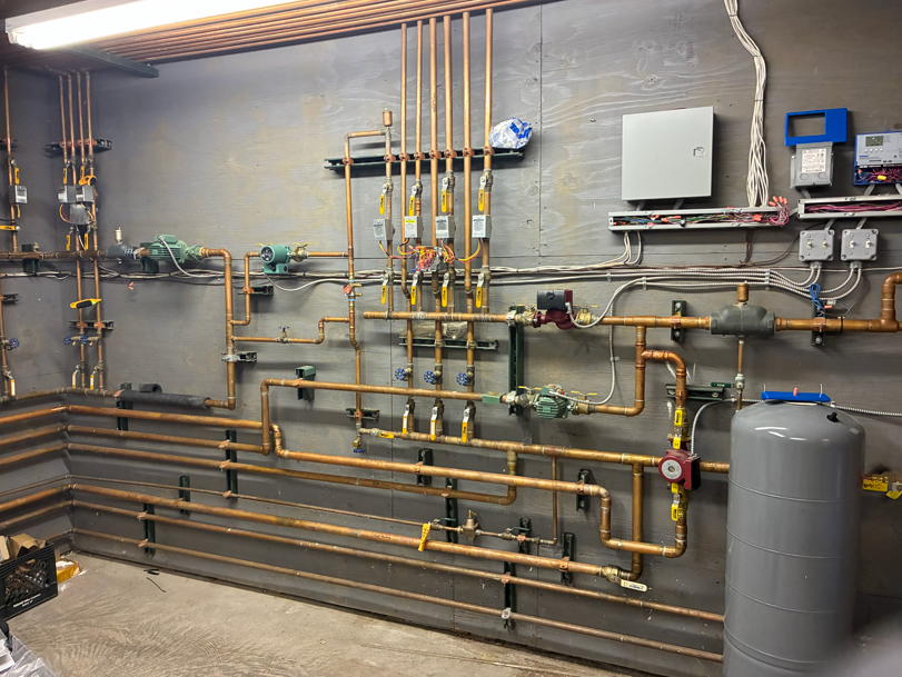 Industrial utility room with exposed copper pipes, valves, gauges, and a large gray tank on the right.