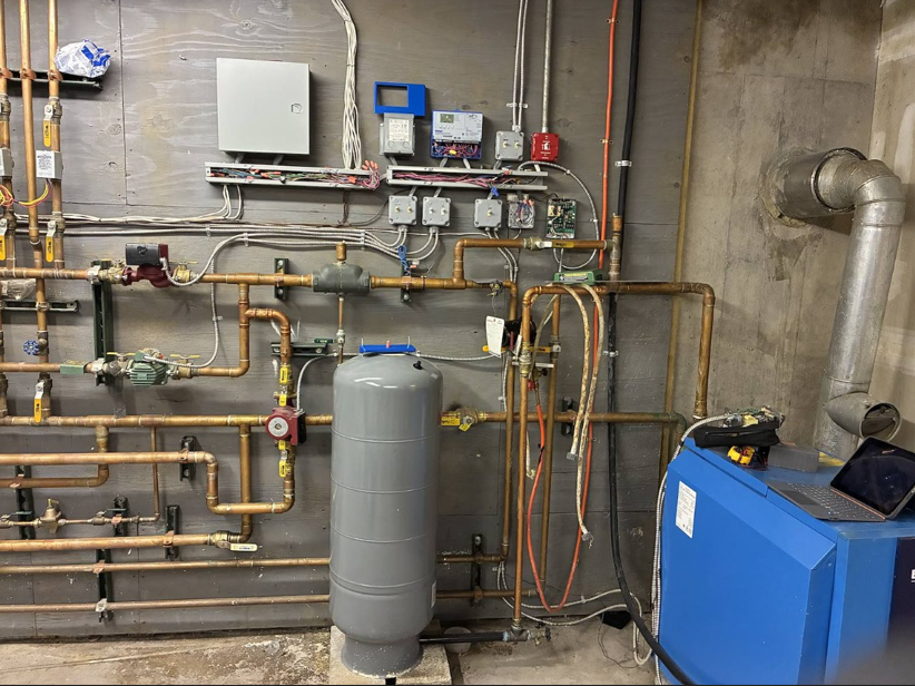 Industrial boiler room with pipes, valves, and a blue heating unit beside a gray tank.