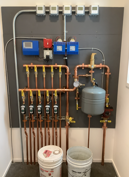 Wall-mounted plumbing manifold with gauges, valves, pipes, and two buckets beneath it.