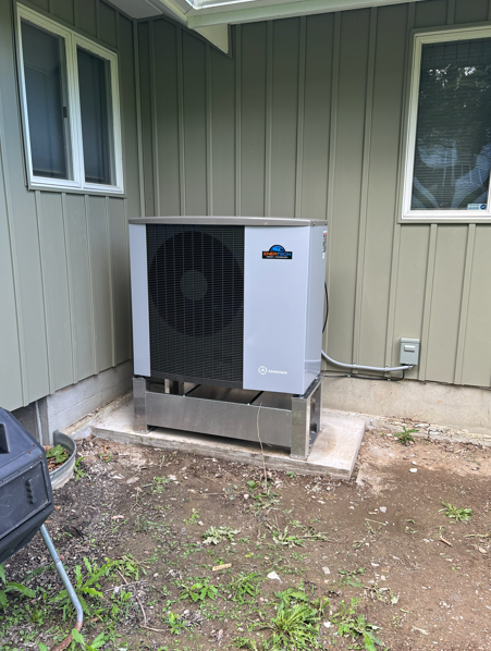 Outdoor HVAC unit beside a house wall, with windows and dirt yard nearby.