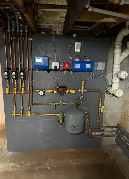 Wall-mounted plumbing and gas control panel with pipes, meters, and valves in a basement utility area