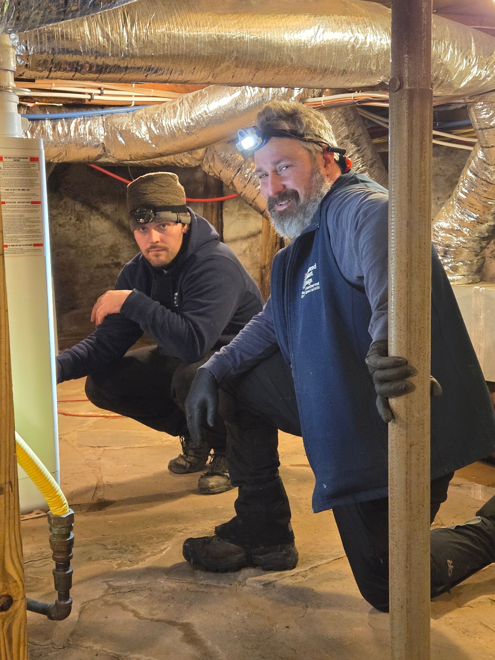 Two people crouch in a crawlspace with exposed ducts and pipes, inspecting the area.