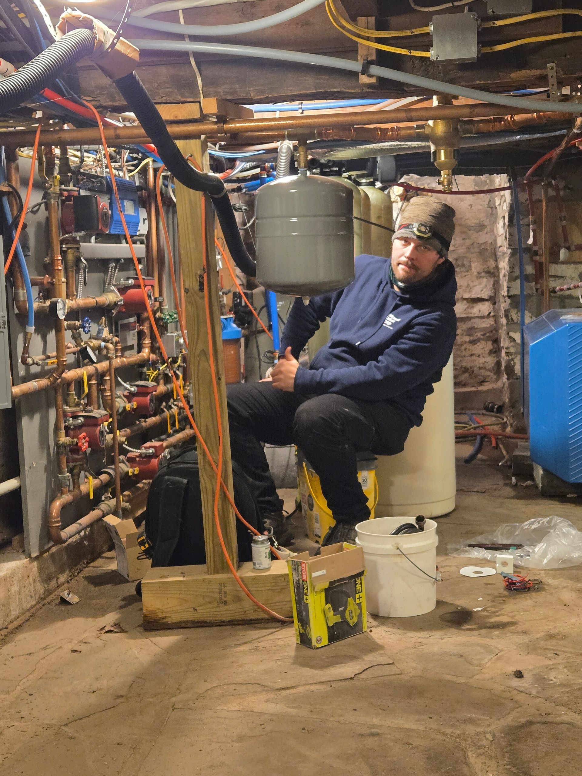 Person sitting in a cluttered basement workshop, surrounded by pipes, tools, and buckets.