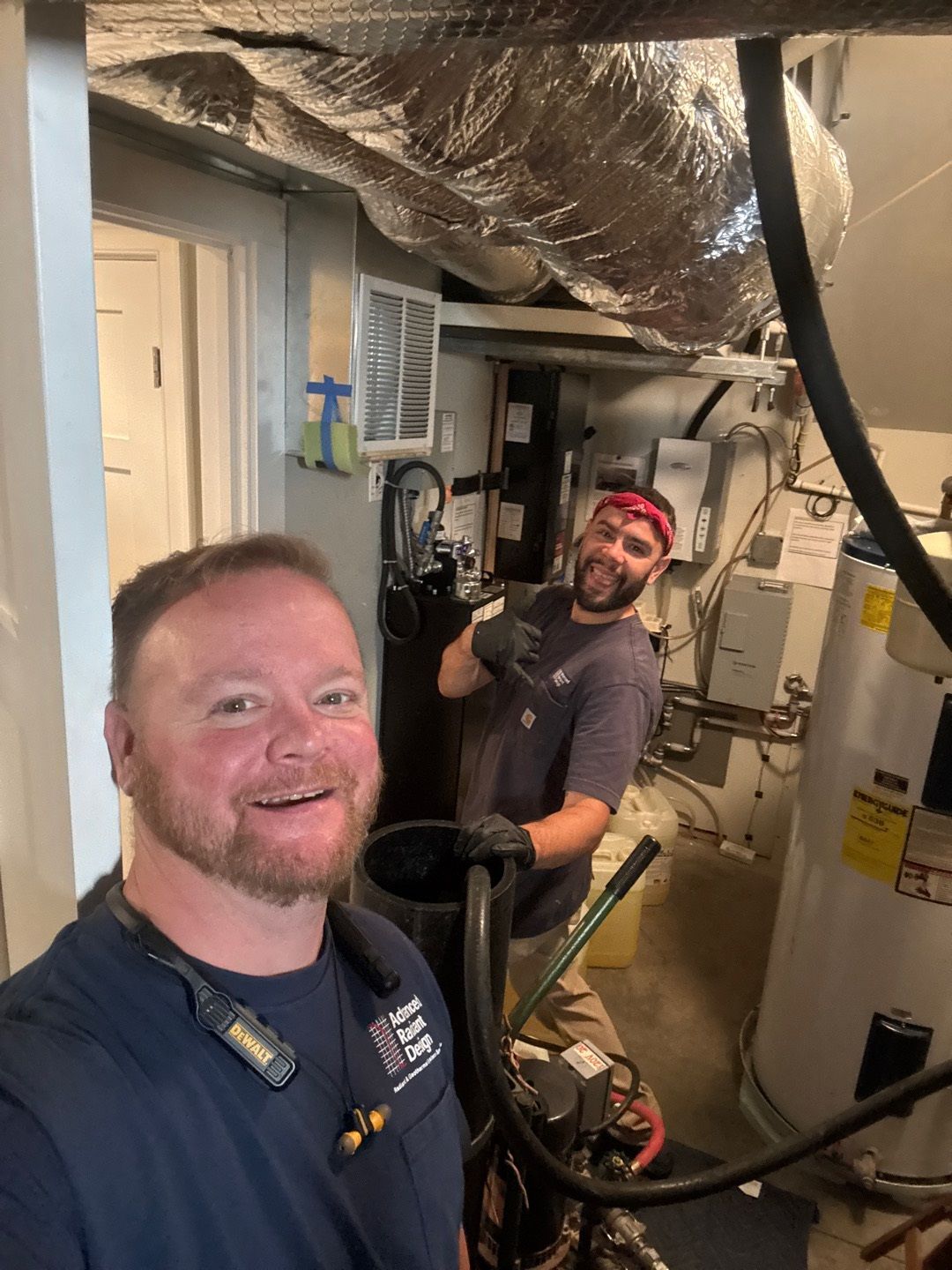Two workers in a mechanical room, one smiling while holding a cable overhead amid pipes and equipment.