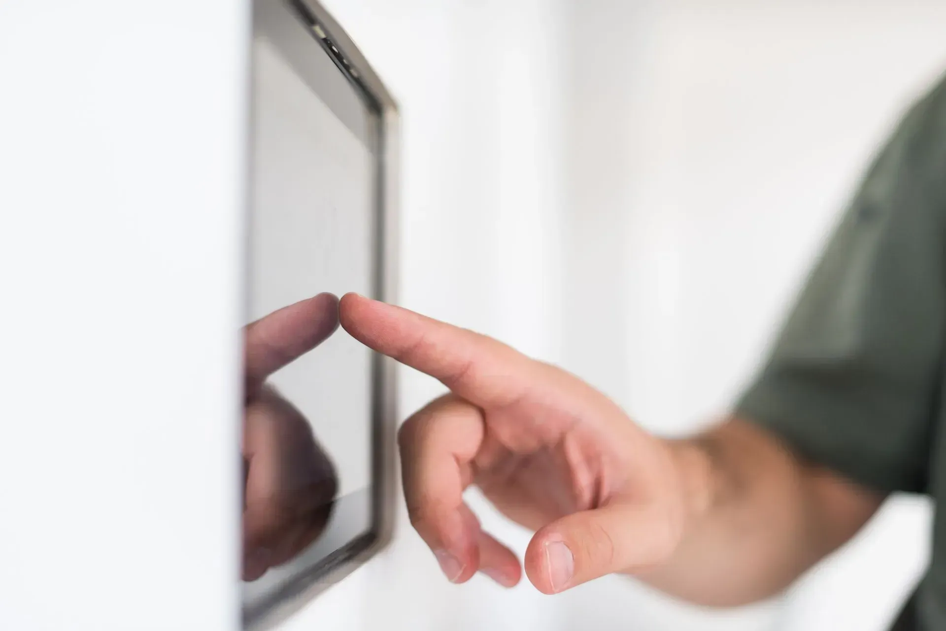 Finger touching a touchscreen beside a white wall, with a blurred reflection on the glass.