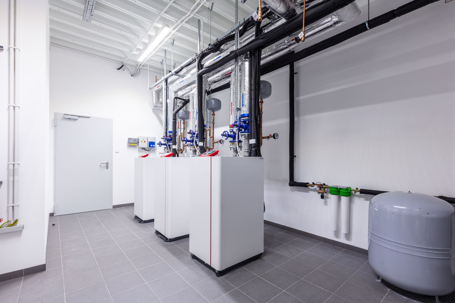 Industrial equipment room with white machinery, pipes, and a gray storage tank on tiled floor