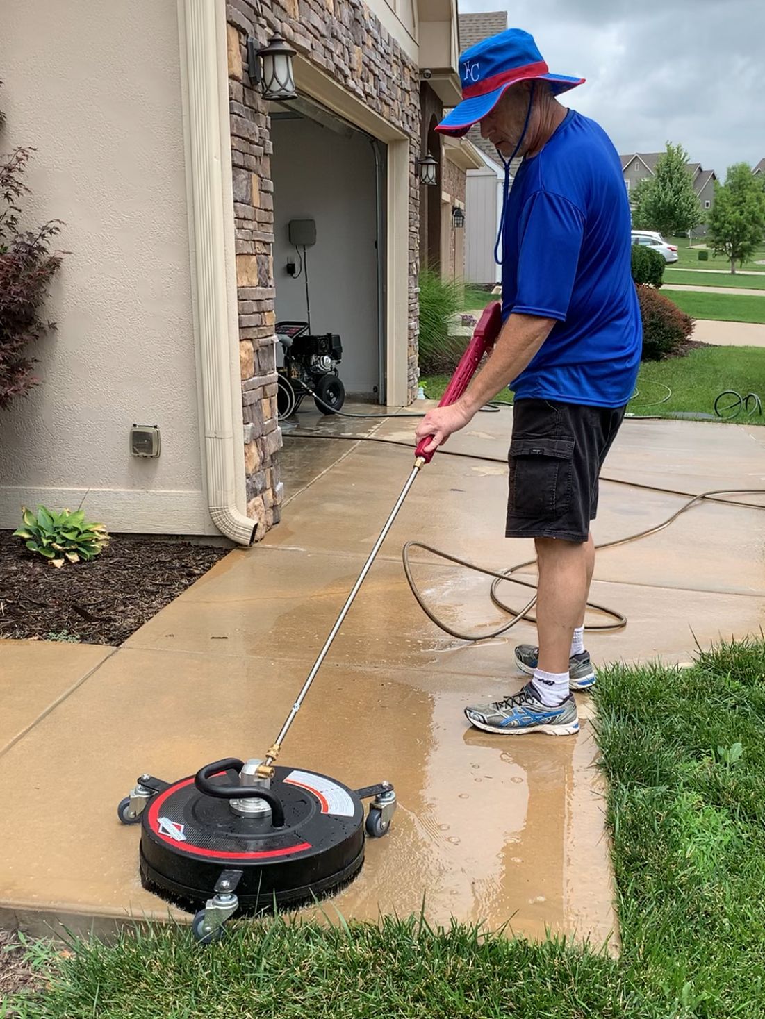 Person power washing a concrete walkway in front of a garage, using a surface cleaner.