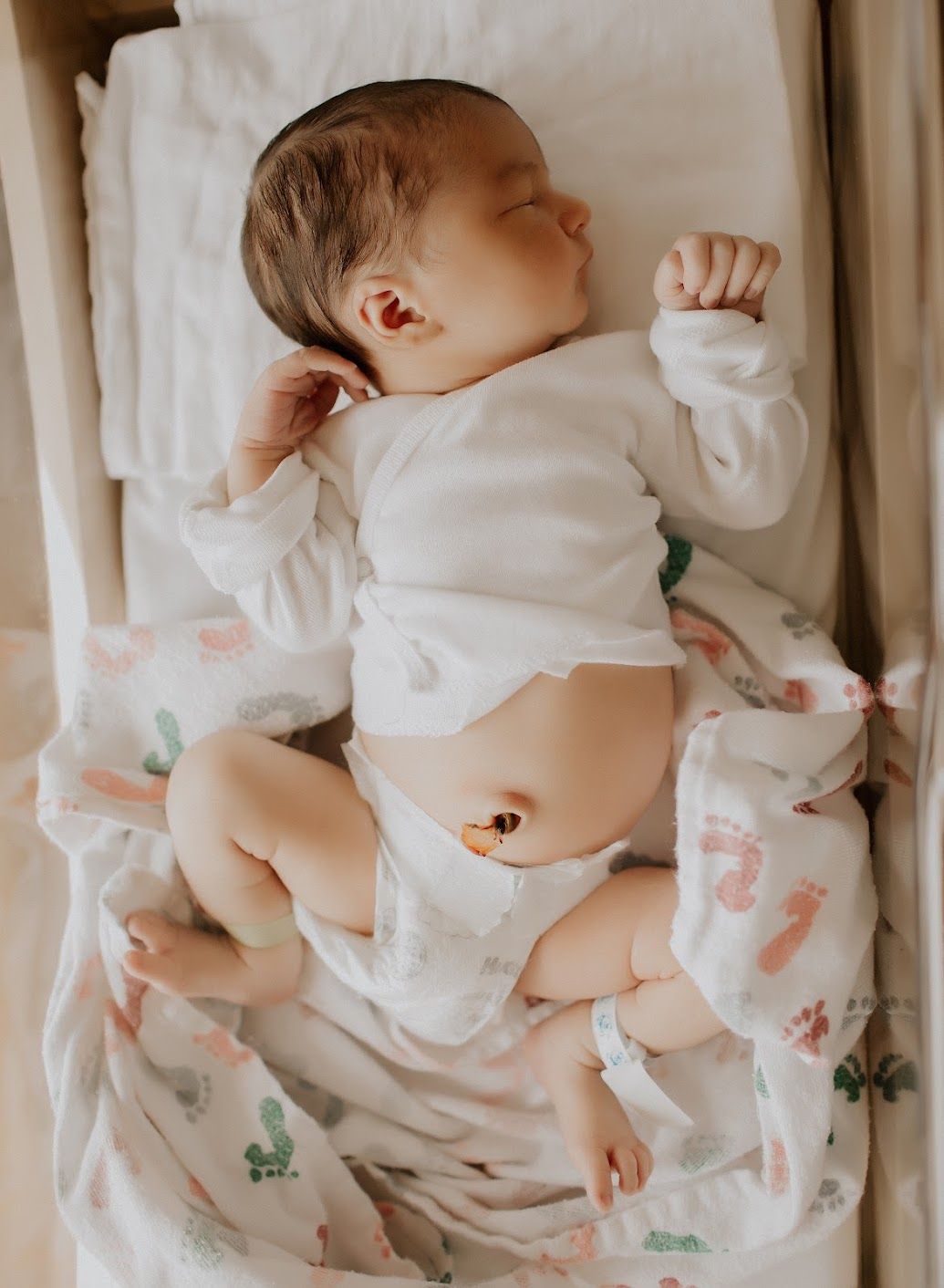 Newborn sleeping in a crib, wearing a white onesie. Light skin, small hands, umbilical cord visible.