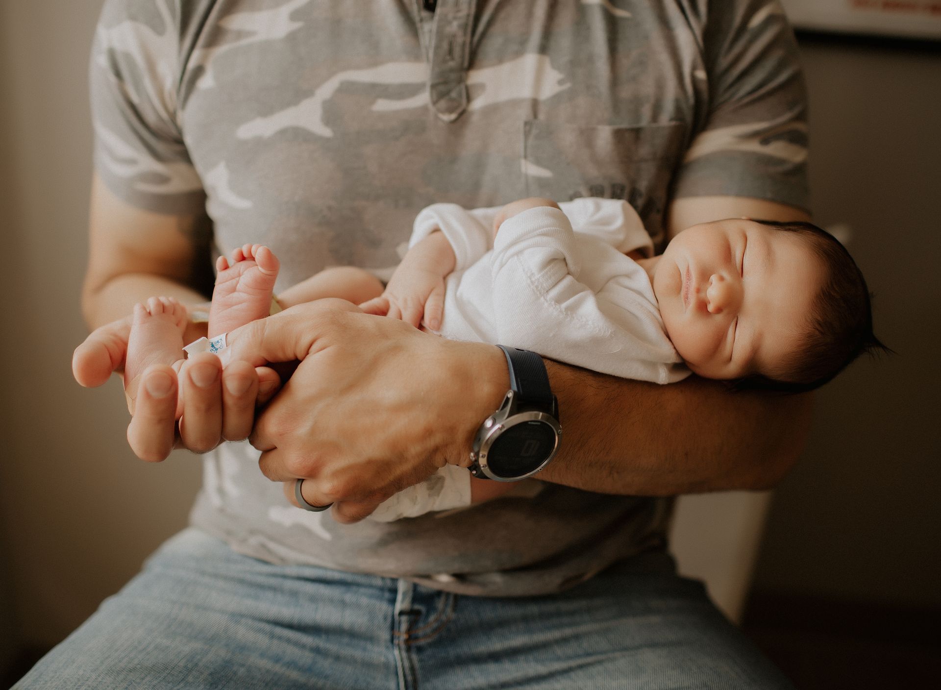Person holding a sleeping baby in arms. Both are indoors. The adult wears a camouflage shirt and jeans.