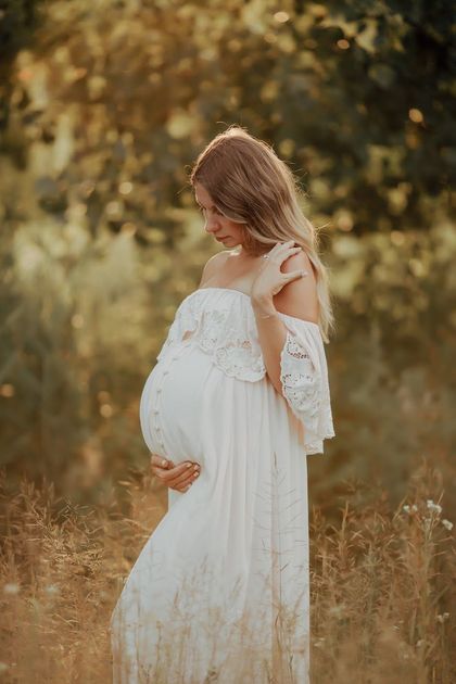 A pregnant woman in a white dress is standing in a field holding her belly during a maternity photography session