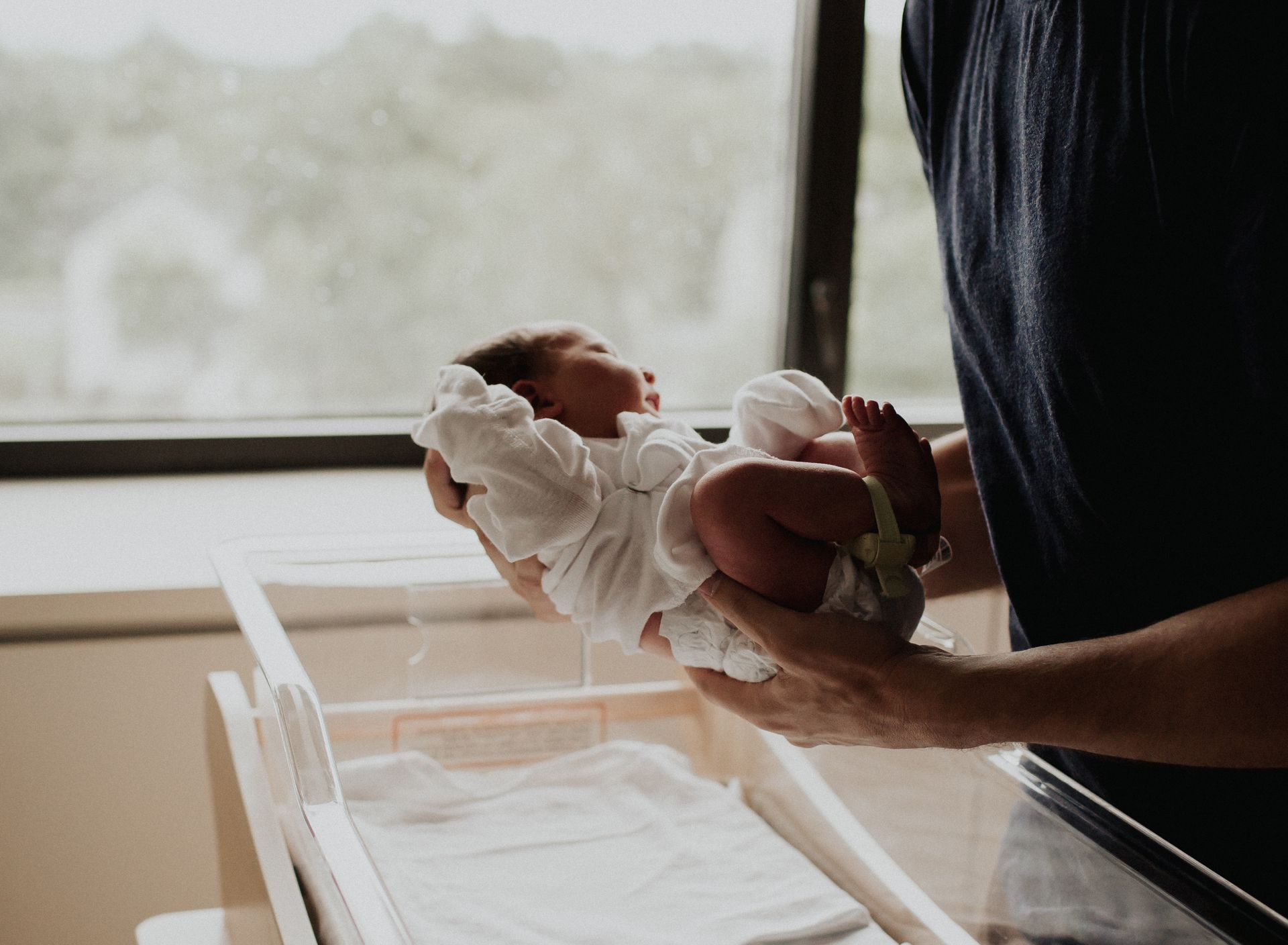 Newborn in white onesie held by person near a hospital bassinet and window.