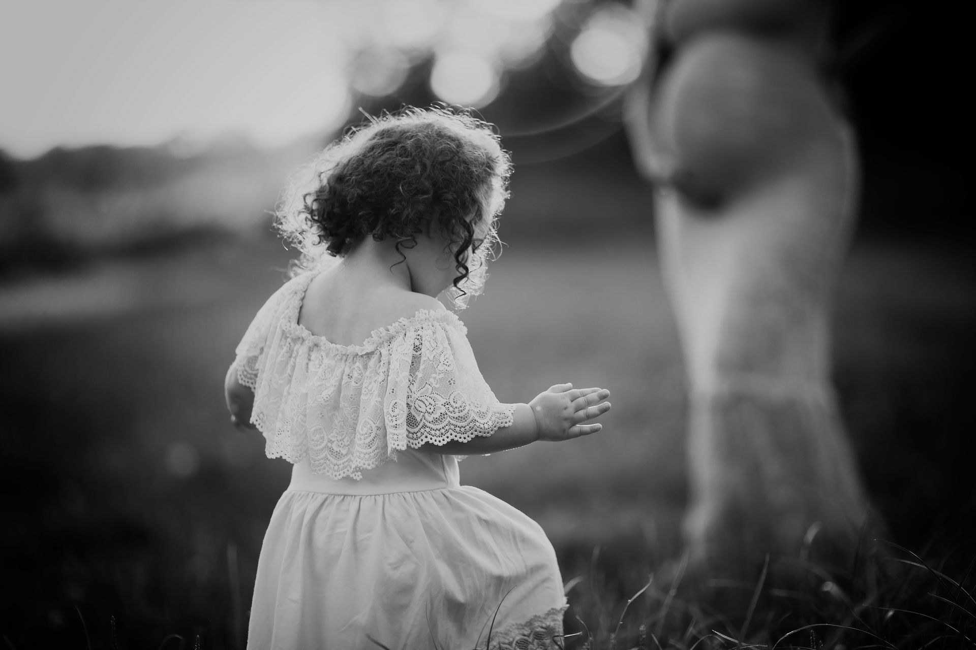 Child in a lace dress reaches toward a pregnant person in a field at sunset.