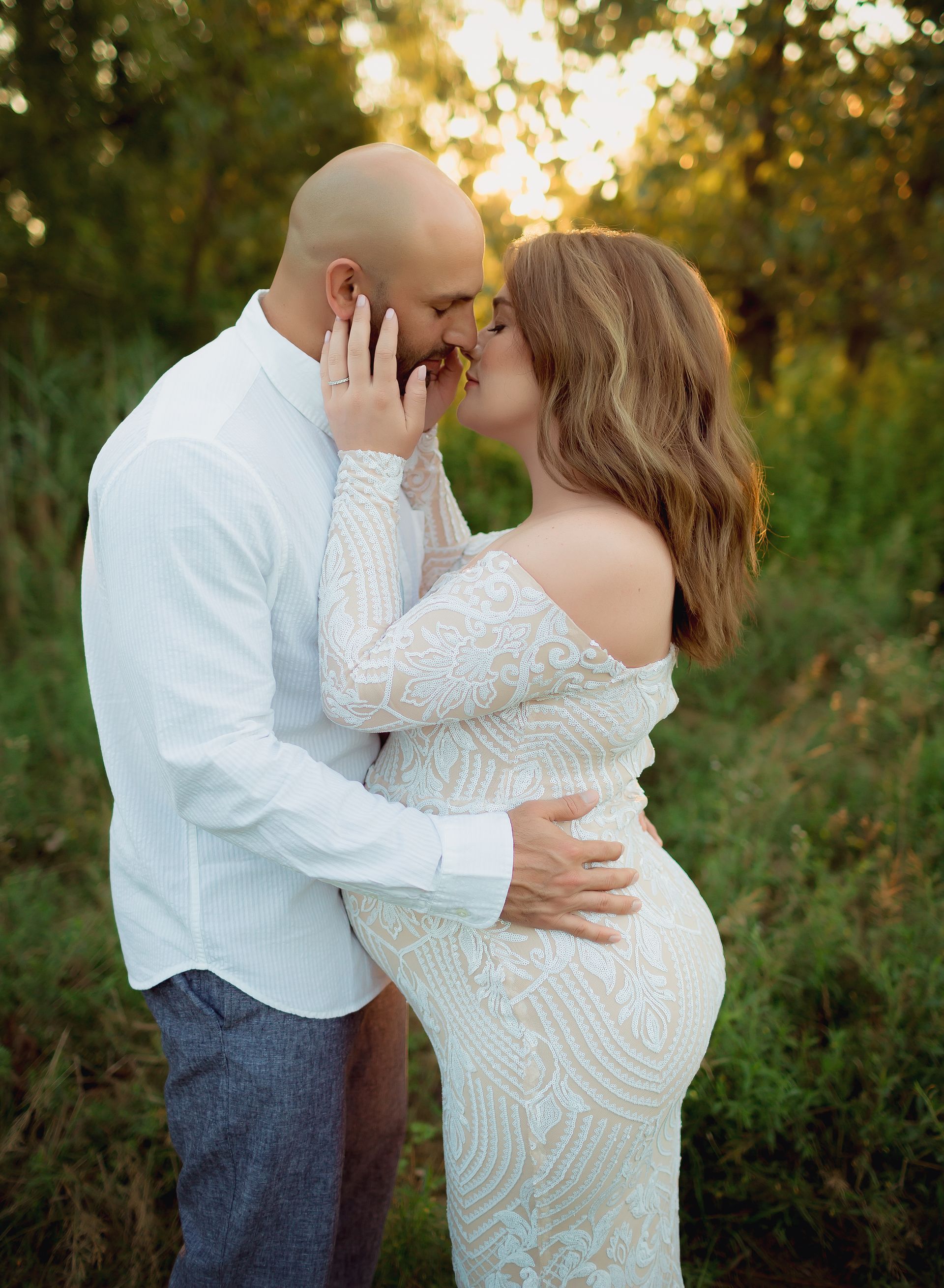 Pregnant woman and partner embrace and kiss in a field at sunset. She wears a white gown, he wears a white shirt.