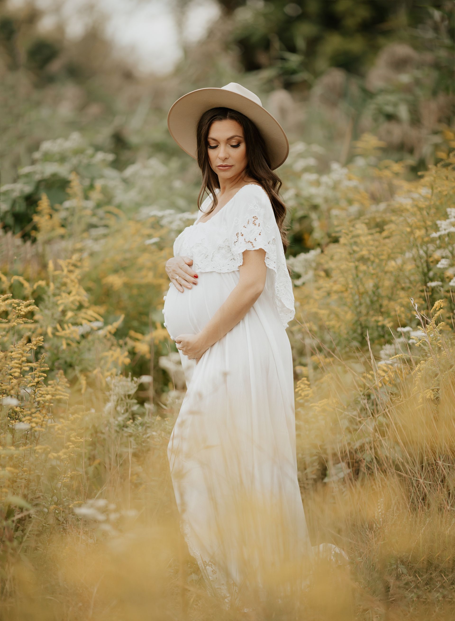 Pregnant person wearing a white dress and hat, holding belly in a field of yellow flowers.