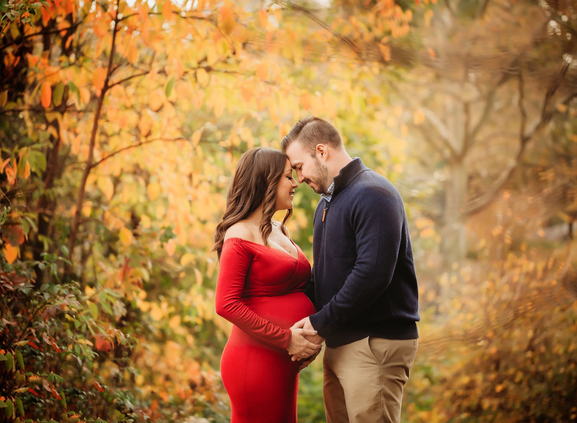 Pregnant woman in red dress with man, embracing outdoors with fall foliage.