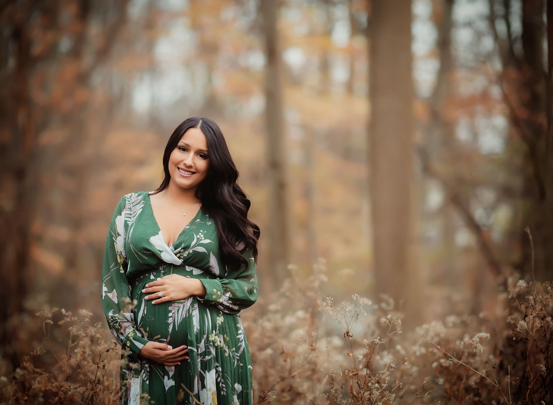 Pregnant woman in green floral dress, smiling, touching her belly, standing in a wooded area with fall foliage.