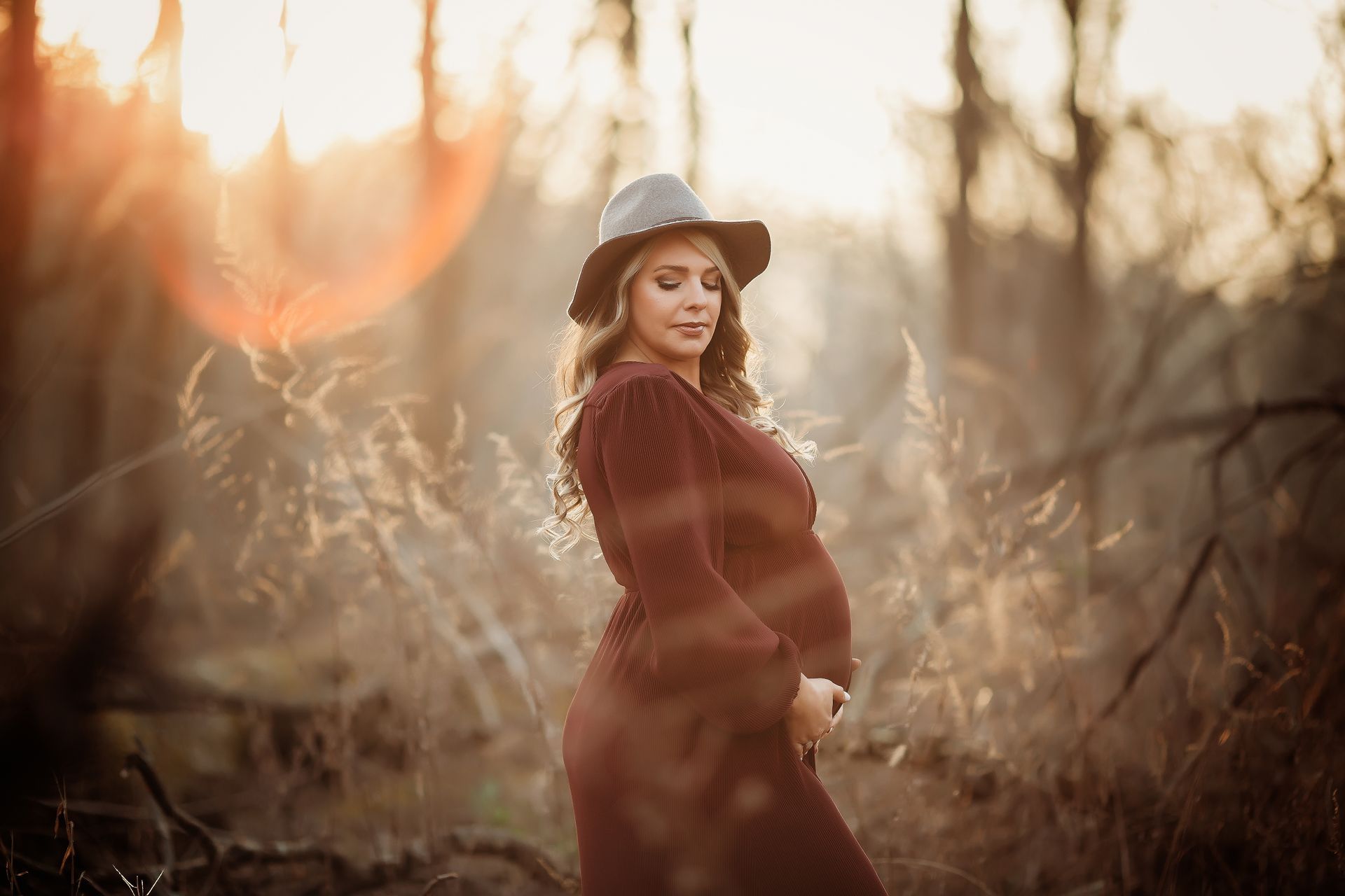 Pregnant person wearing a hat and long dress, holding her belly, standing outdoors with golden sunlight.