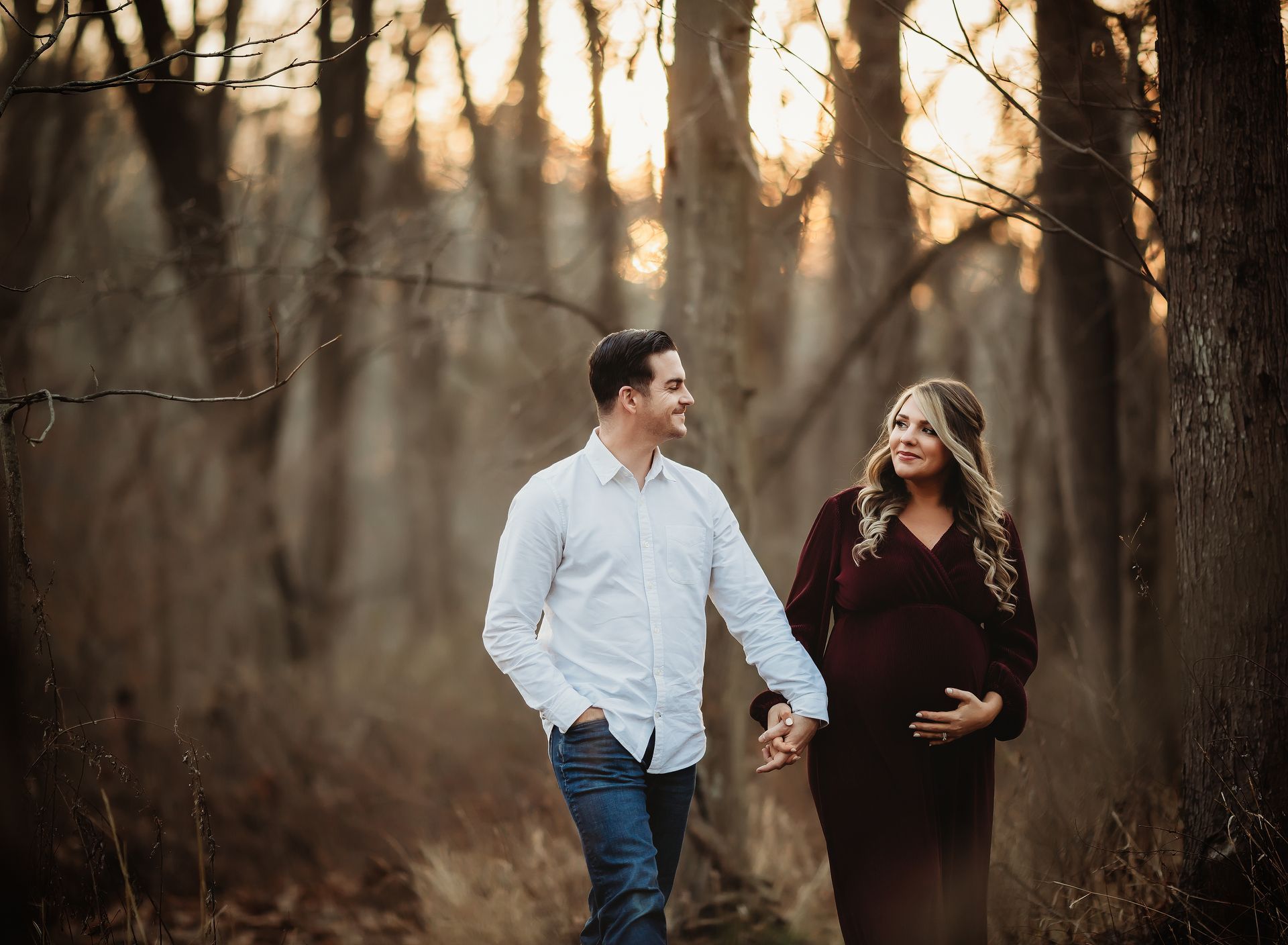 Couple holding hands, walking in a wooded area, woman pregnant, man smiling, sunset background.