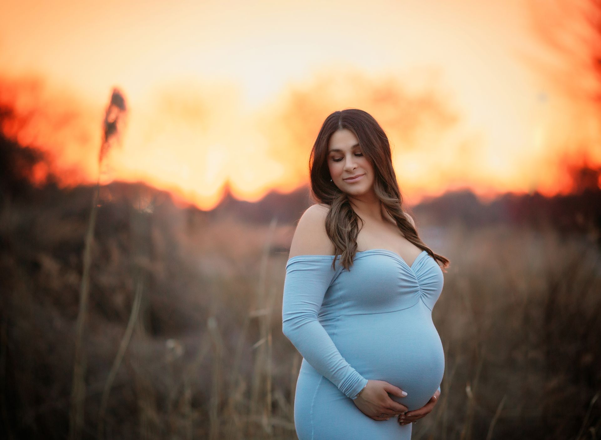 Pregnant person in a light blue dress cradles belly. Sunset backdrop over field.