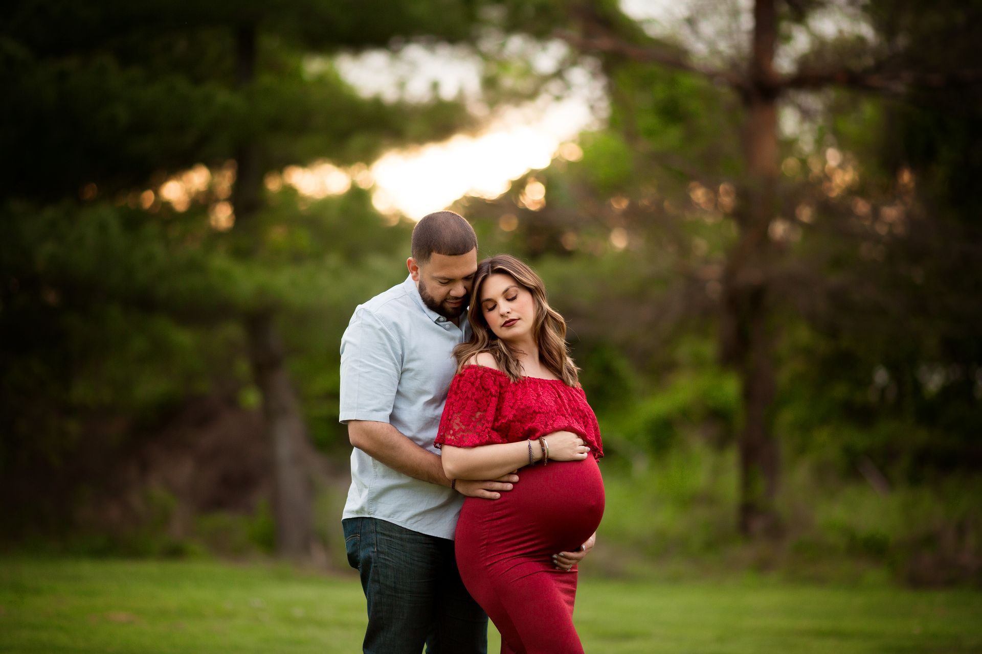 Pregnant woman in red dress with partner in park, sunset in background.