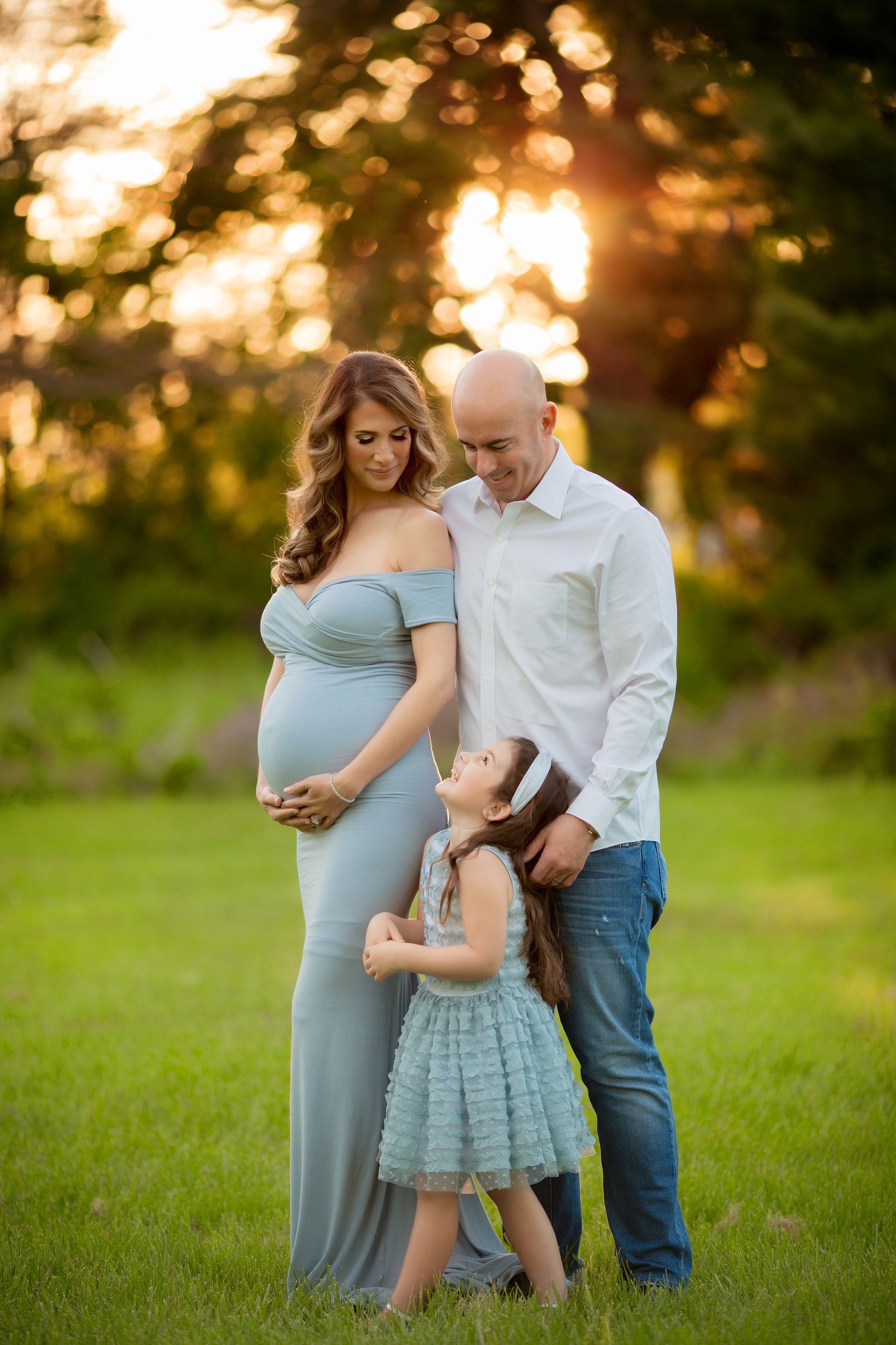 Pregnant woman and family outdoors, touching her belly. Sunlight backlights the scene.