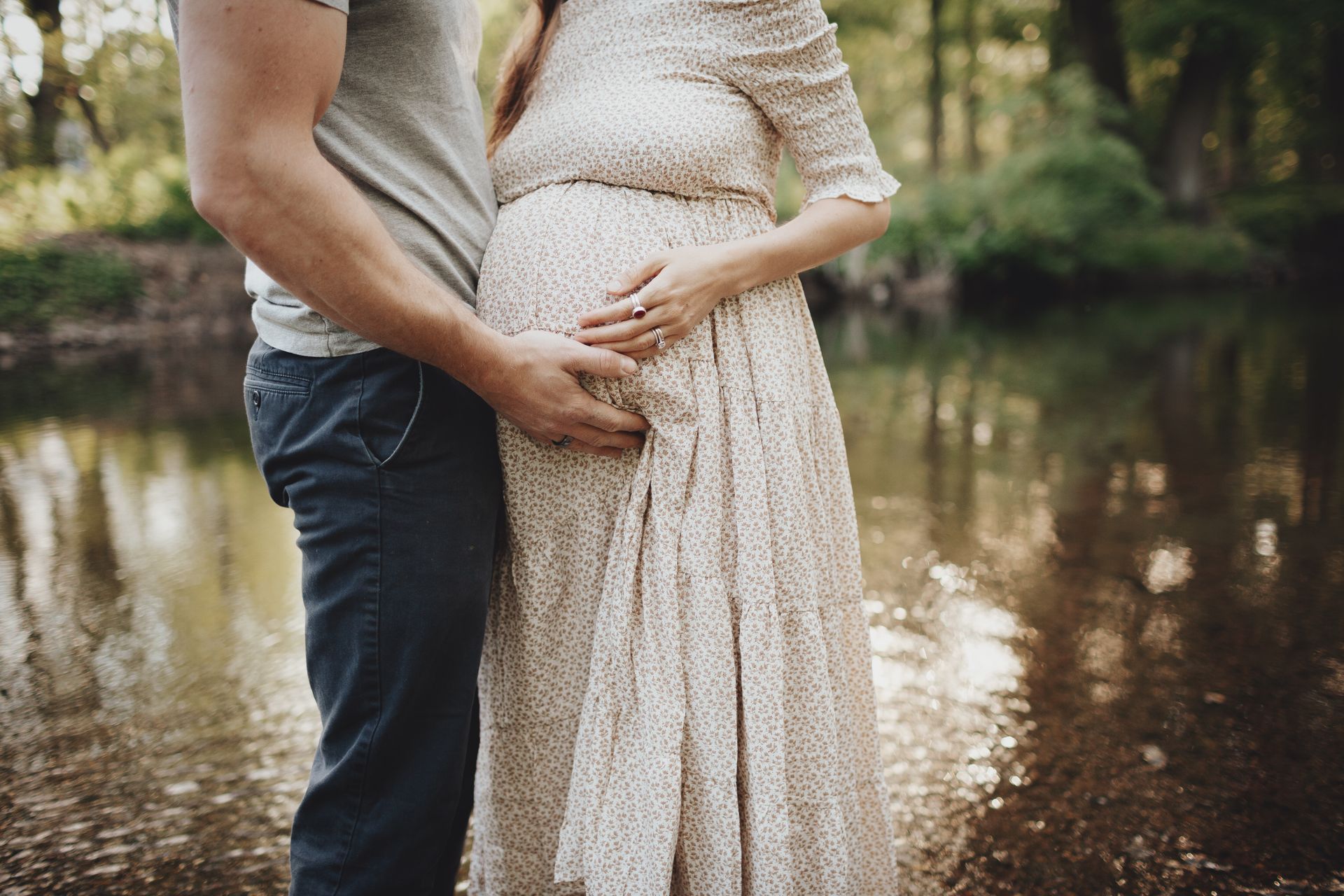 Pregnant person and partner touching baby bump near a creek.
