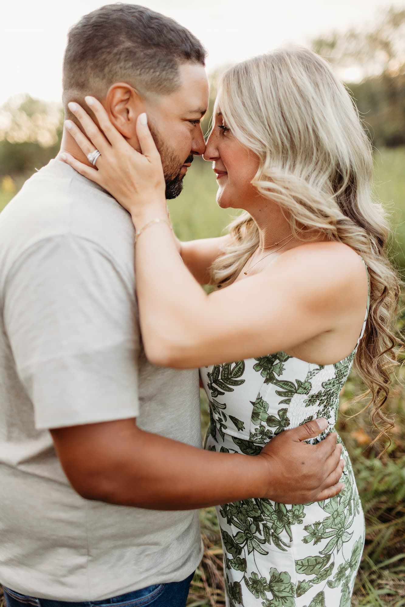 Couple embraces in a field. Woman in white floral dress, man in tan shirt. Both have arms around each other.