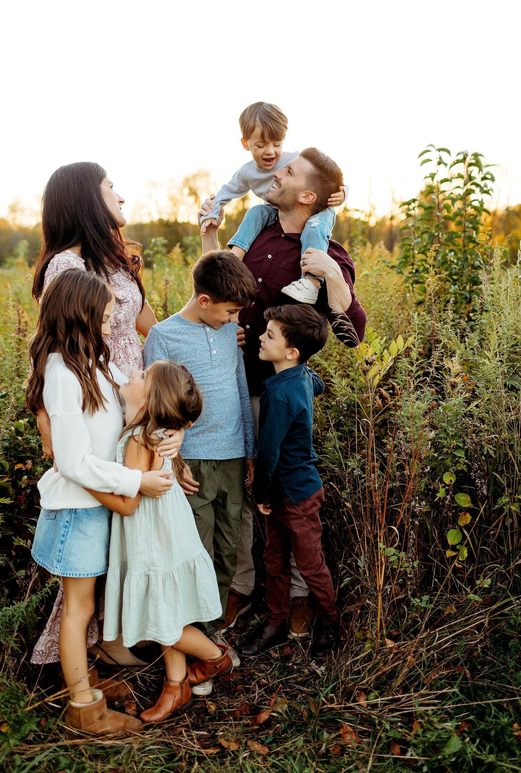 Family of eight smiling in a field; one child on father's shoulders, others hugging and looking toward each other.