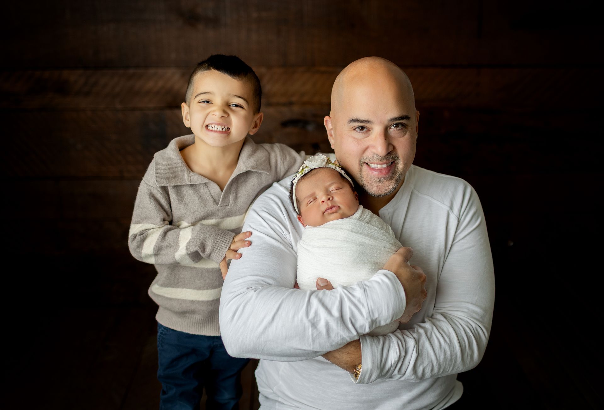 Father holds newborn, smiling. Older child smiles behind them. Dark wood background in Passaic County, NJ.