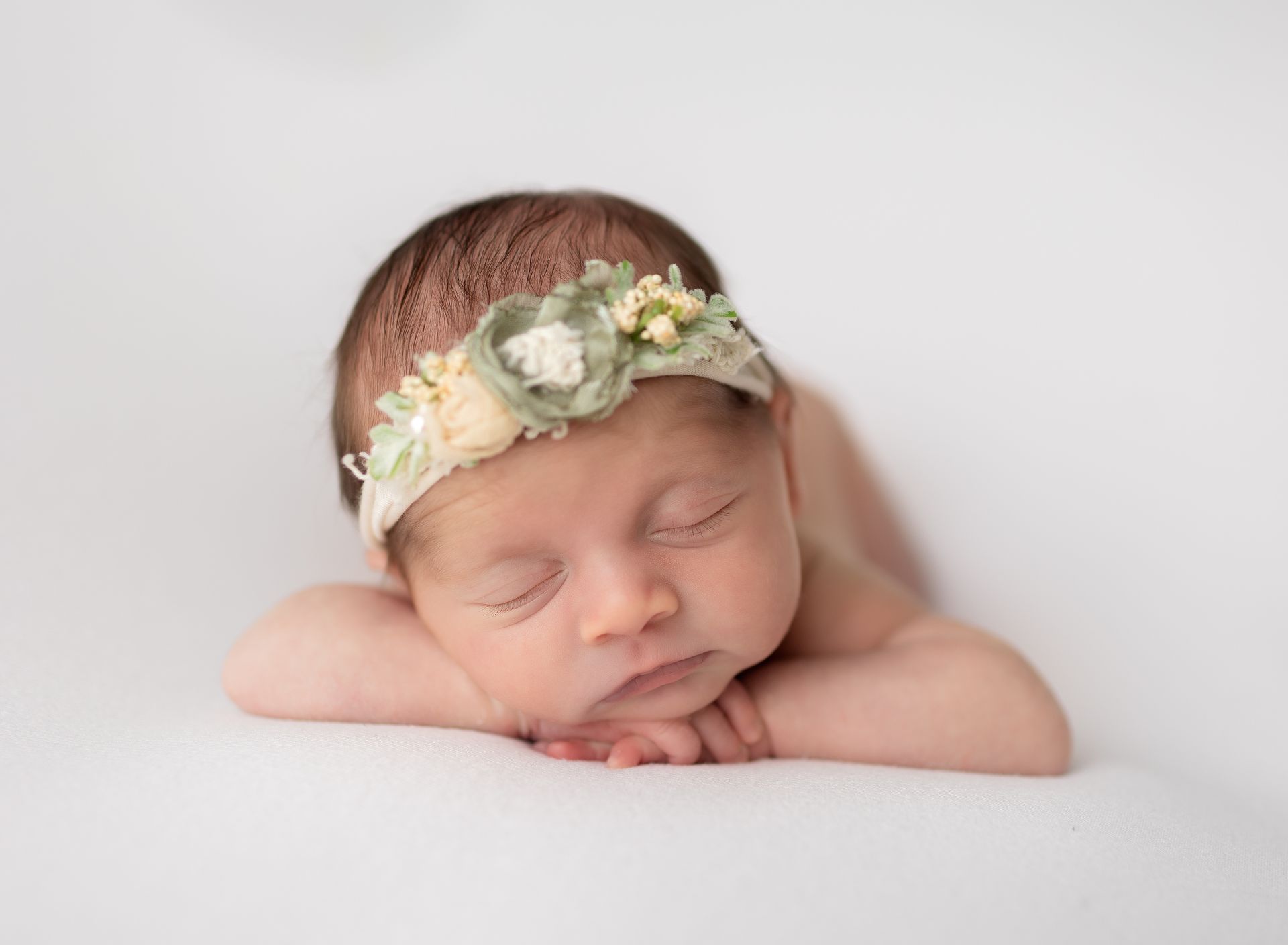 A newborn baby wearing a flower headband is sleeping on a white blanket.