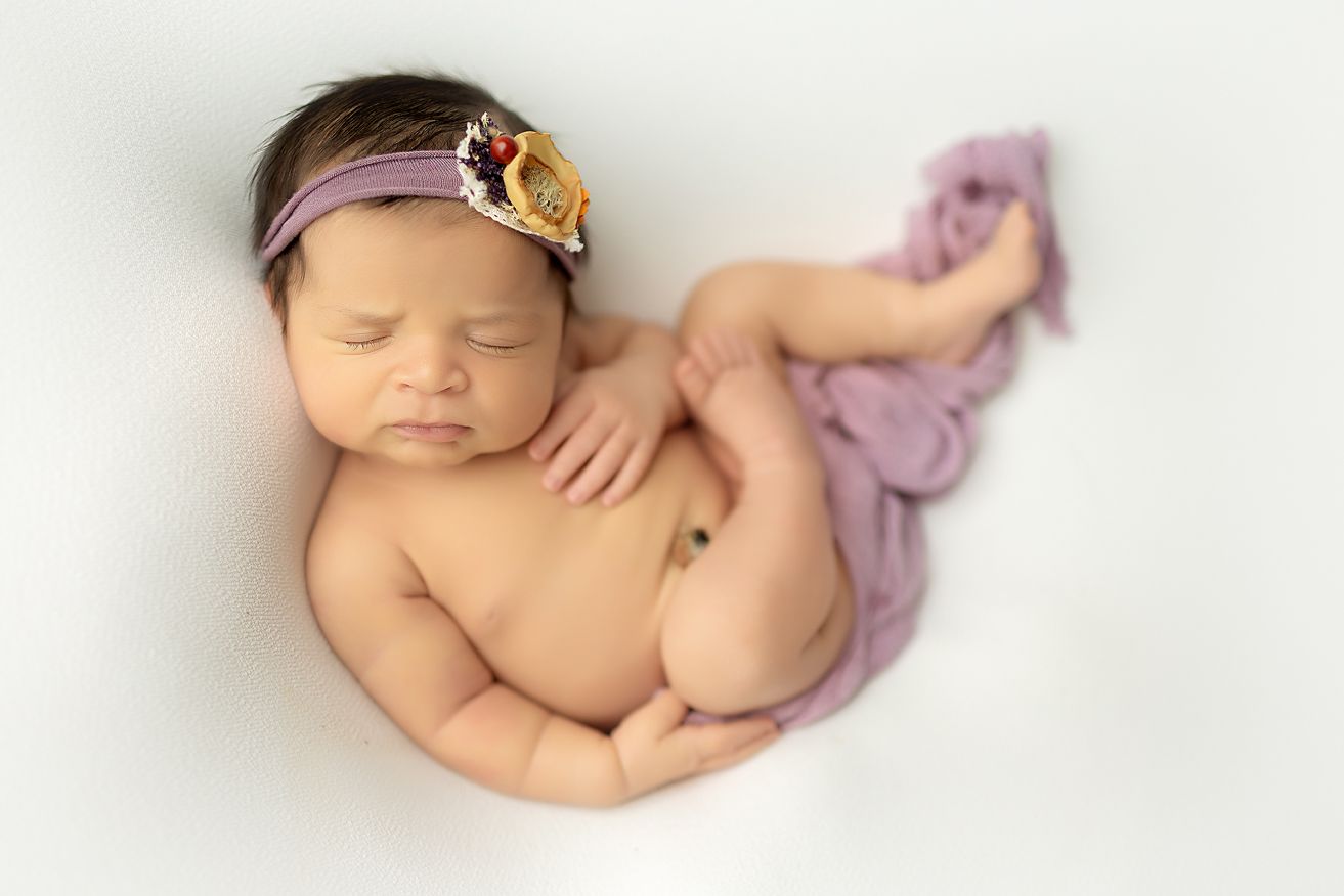 Newborn baby curled up on a white background, wearing a purple headband with a flower.