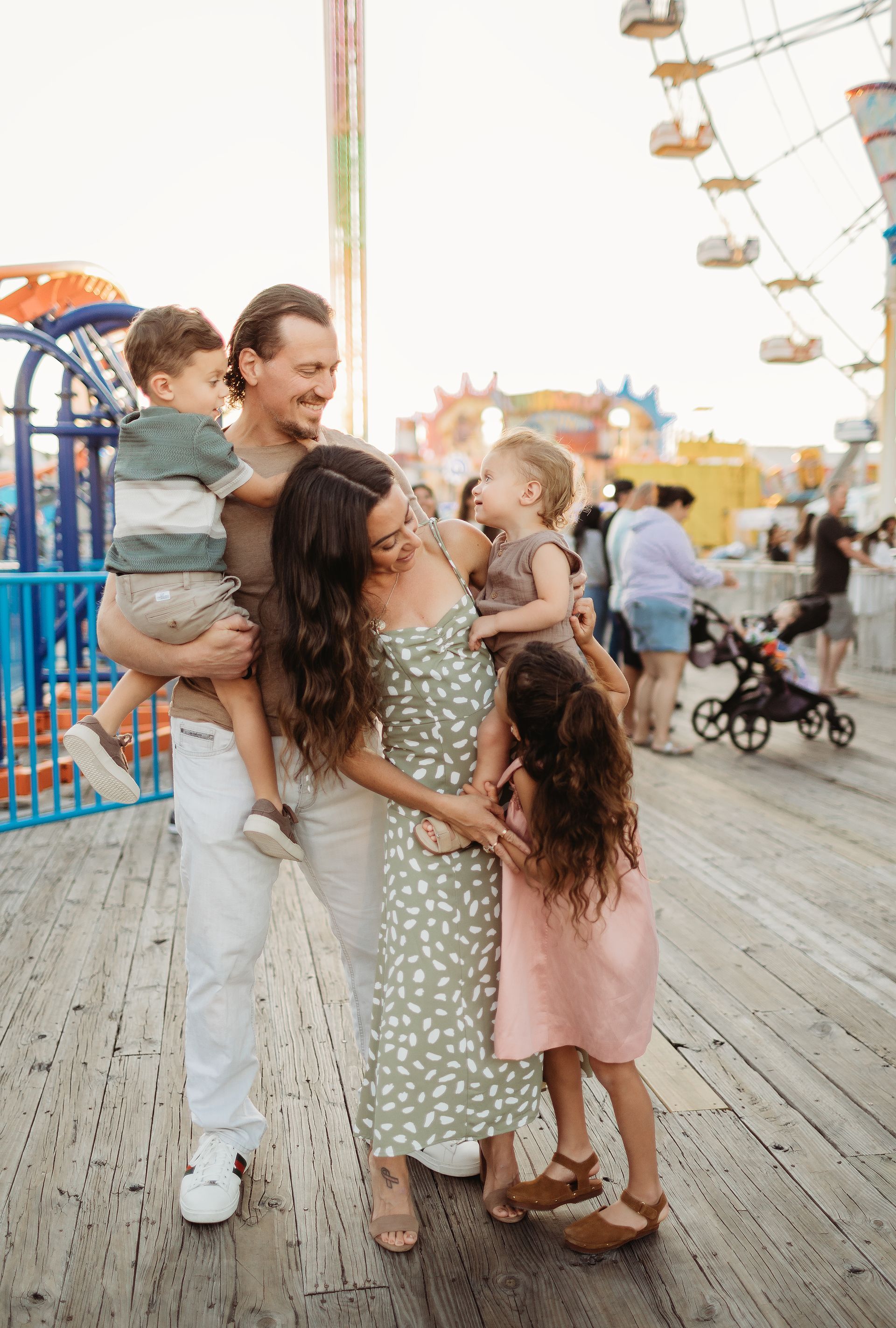 Family of five at an amusement park: father holding a boy, mother hugging a boy, and girl in front of them.