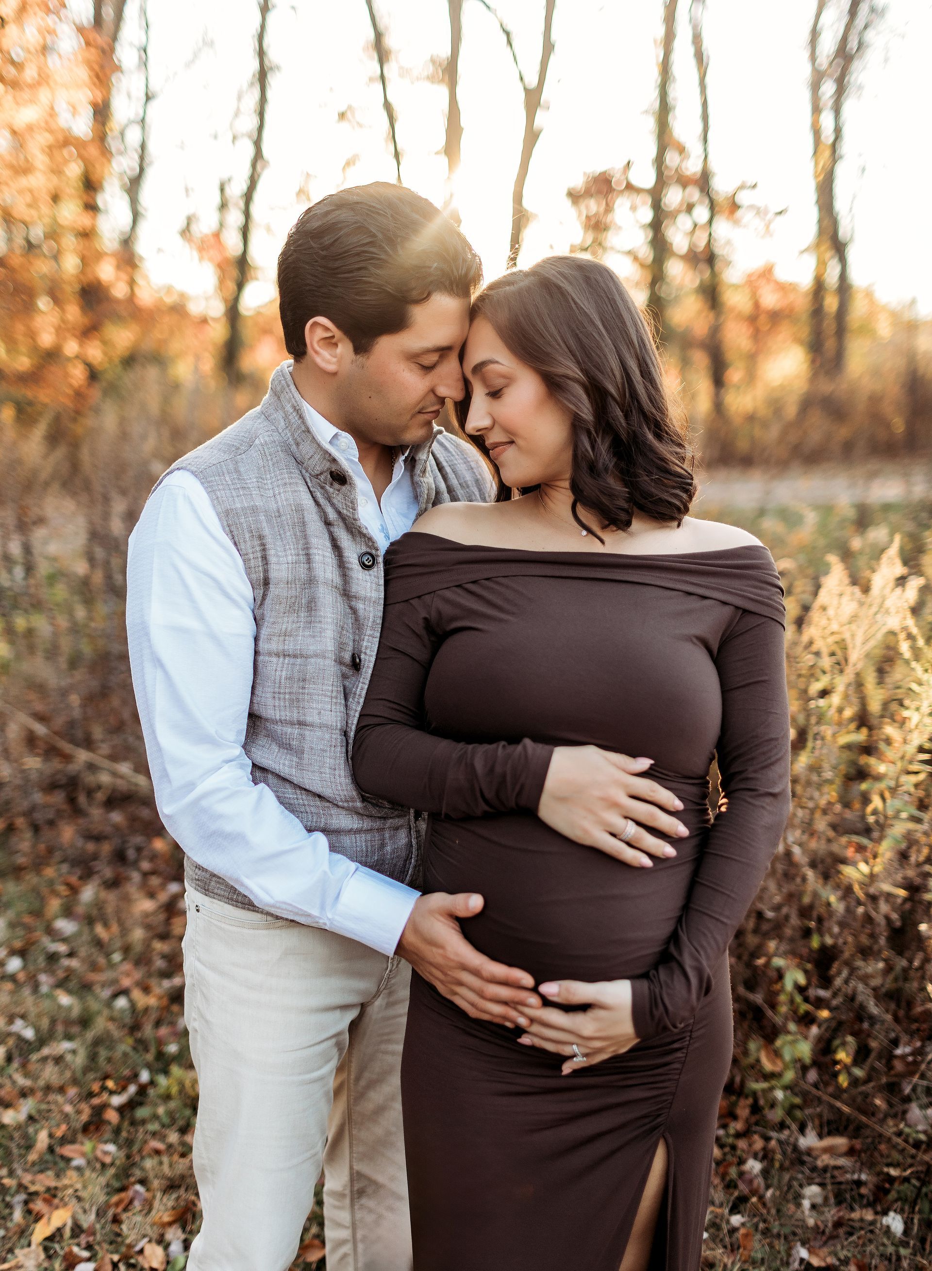 Pregnant woman in brown dress, embracing her partner in a field; autumn colors, touching bellies.