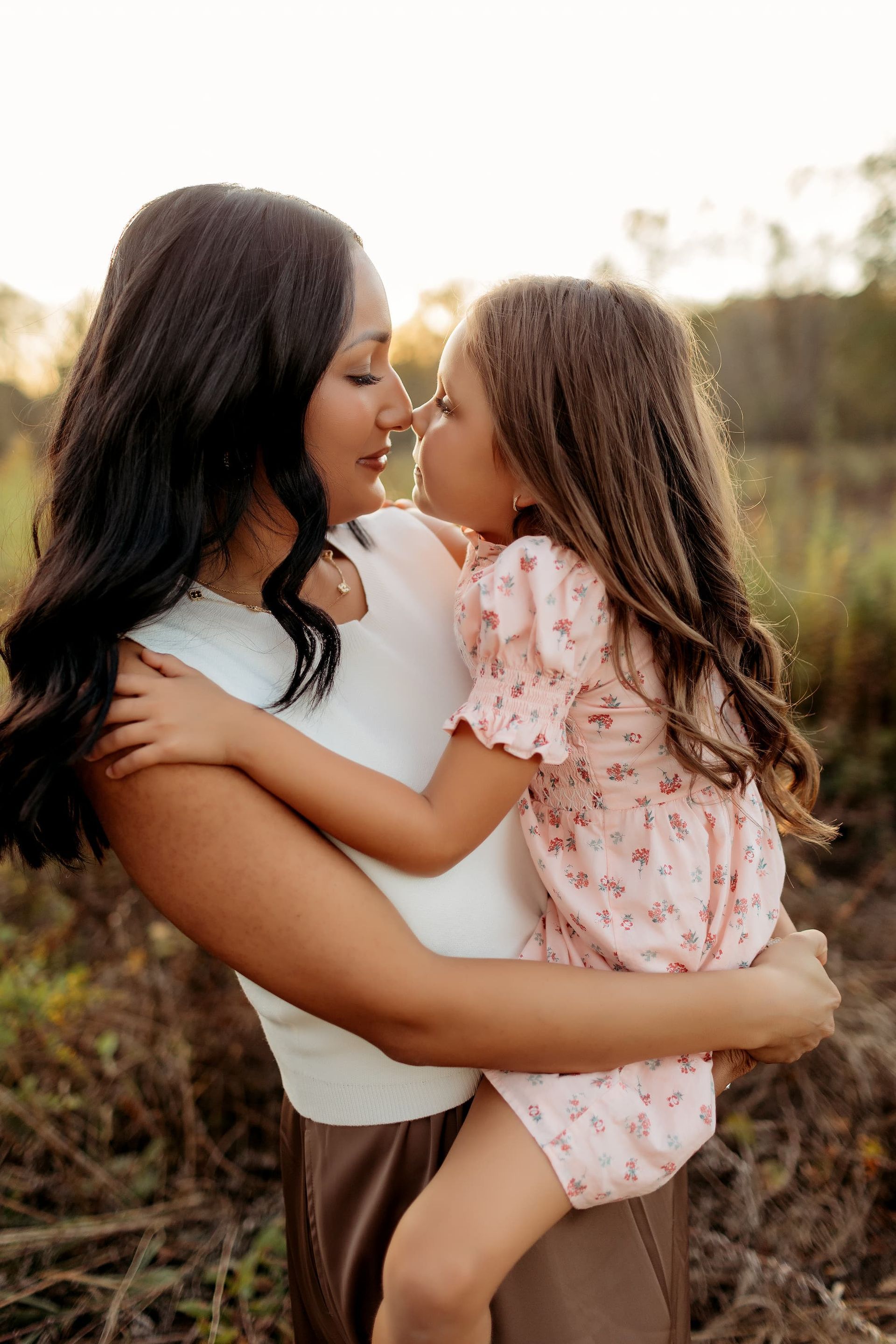 Woman holding child nose-to-nose, smiling in field with golden sunlight.