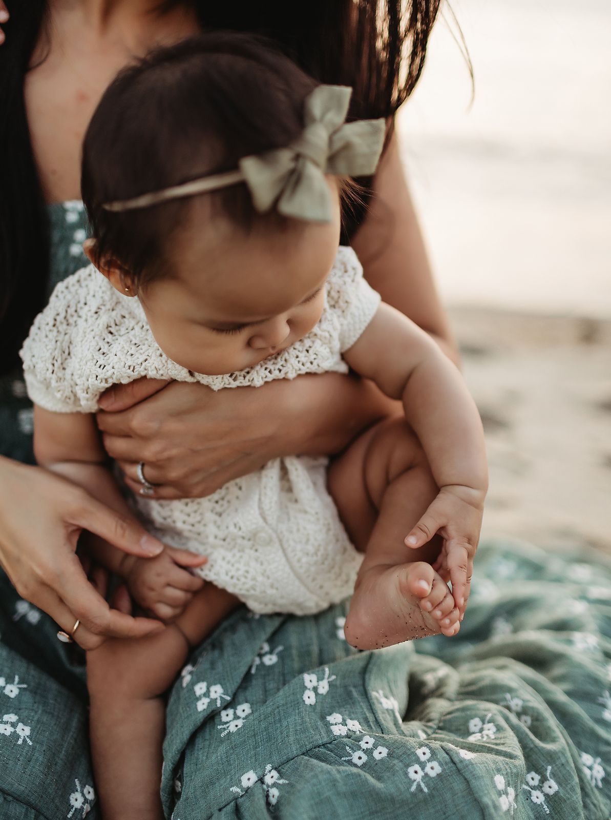 Baby in white lace romper and green bow, held by a person wearing a green floral dress at a beach.