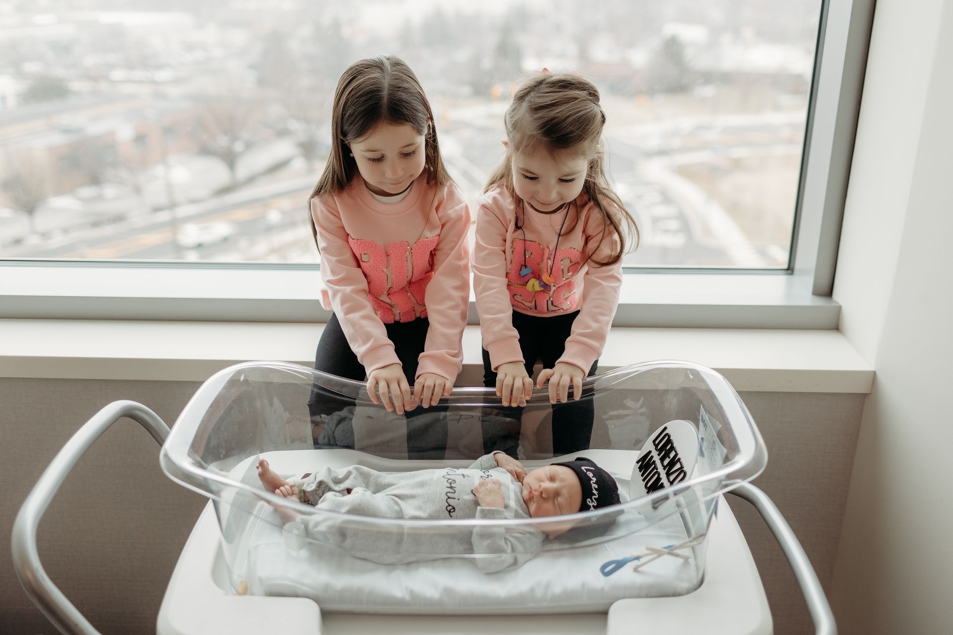 Two young children peer into a bassinet holding a newborn baby. They wear pink tops and black pants by a window.