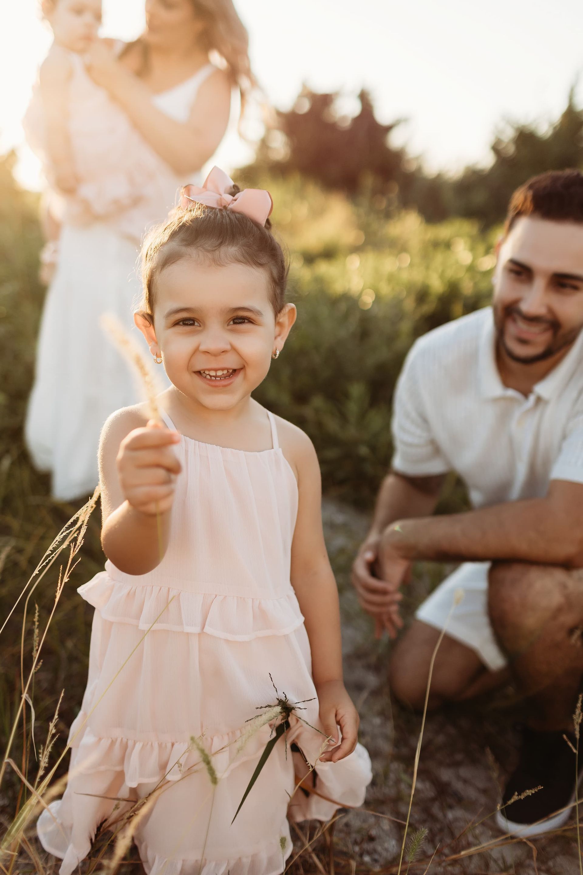 Girl in pink dress holding grass, smiling at the camera, with family in a field.