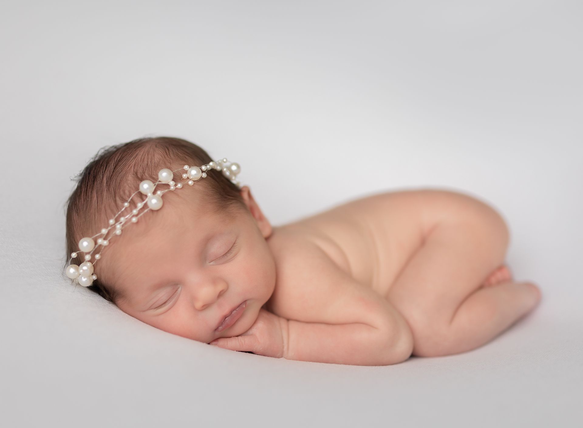 A newborn baby wearing a pearl headband is sleeping on a white blanket.