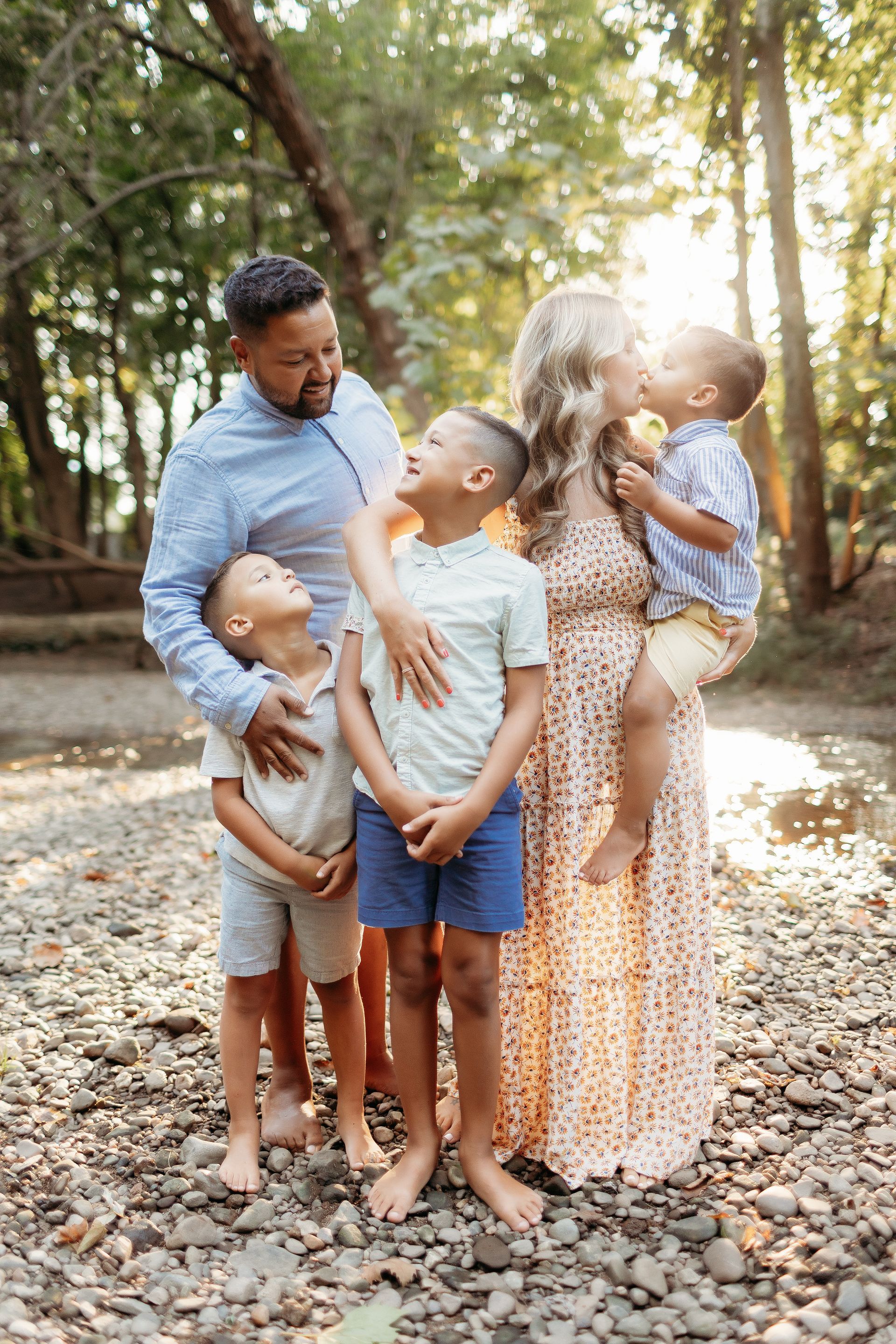 Family of five posing outdoors in a wooded area, smiling, with the mother kissing one son.