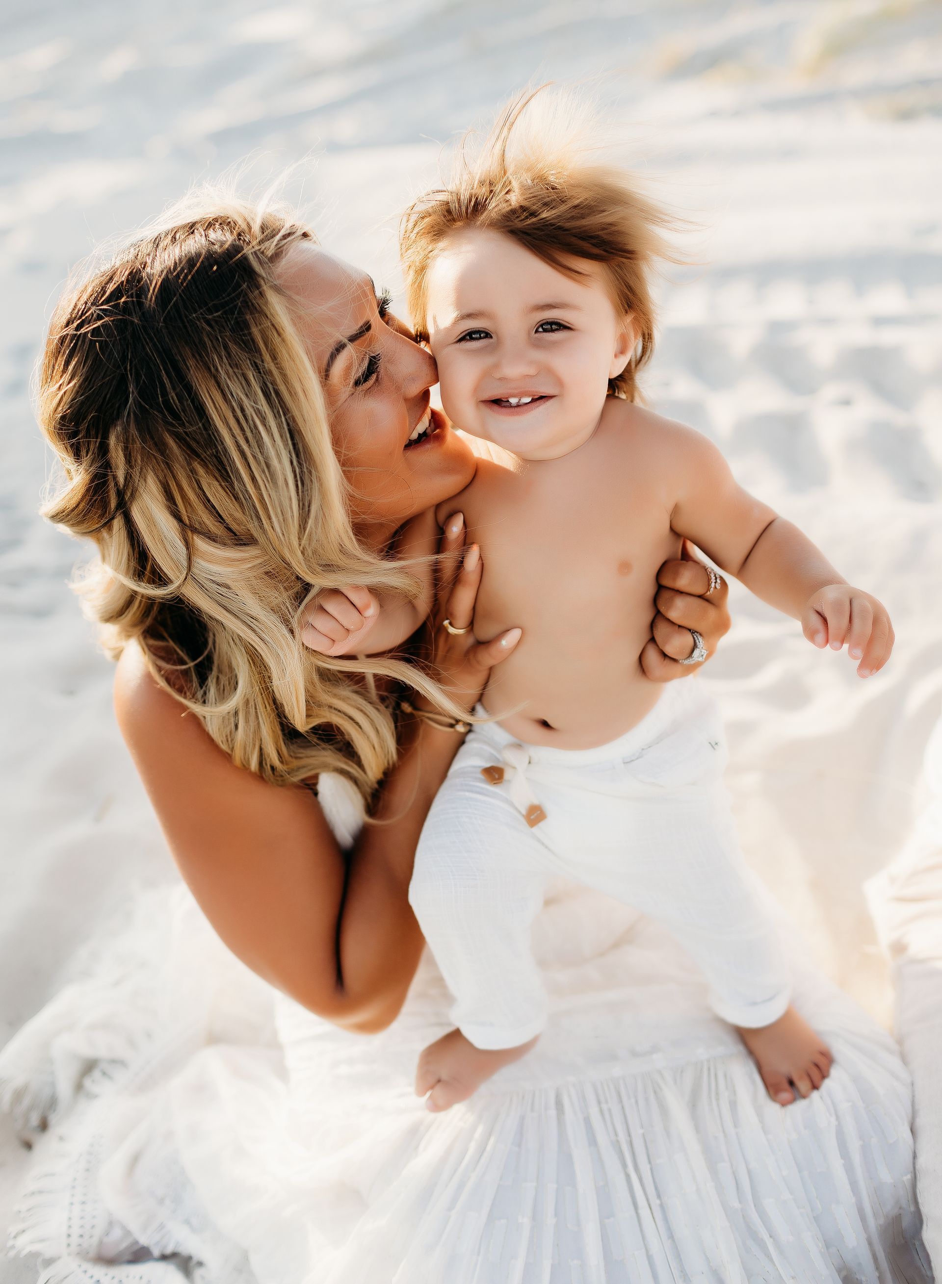 Woman holding and kissing a child on a beach, both smiling. The child is wearing white pants.